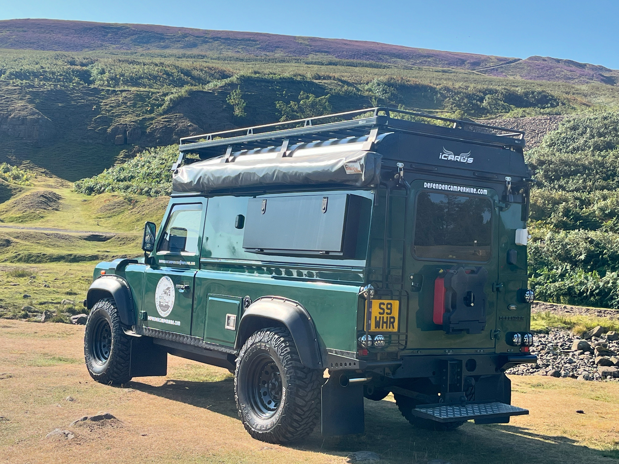 Rear view of a green Land Rover Defender camper in the hills